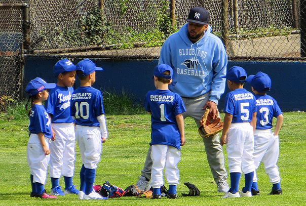 A youth baseball coach in a sweatshirt and Yankees hat talking to his team 