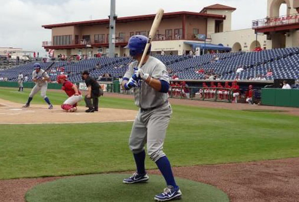 Batter standing in the on deck circle wearing grey jersey with blue helmet and watching pitcher deliver pitch to current batter