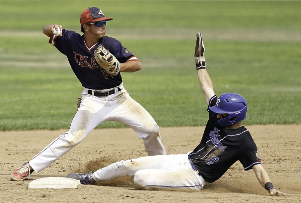 Player in black jersey with purple helmet sliding into second base with arm raised while defender is turning to throw ball towards first base