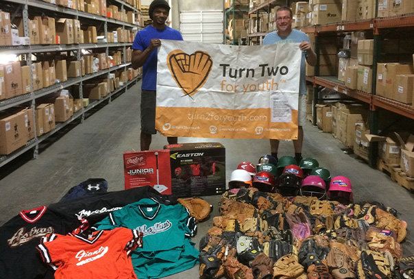 Two men hold a Turn 2 For Youth sign while standing in warehouse in front of a bunch of donated baseball gear and boxes