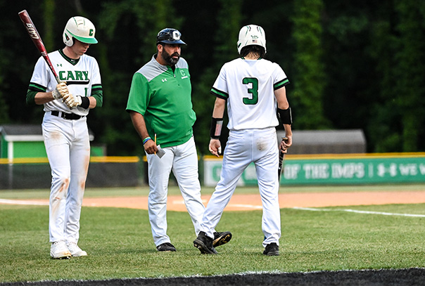 Two Cary High School players, one holding a bat and one holding batting gloves, standing on grass speaking with coach