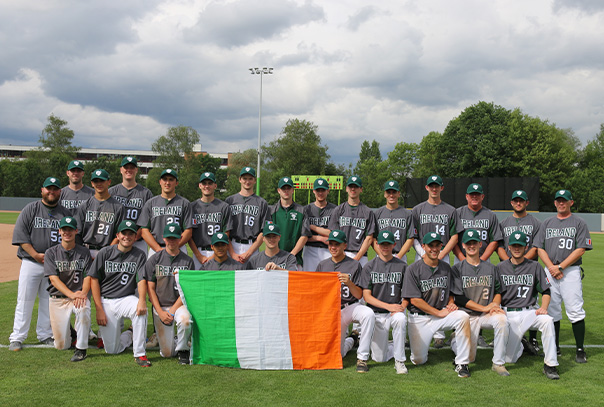 Team Ireland standing on baseball field in grey uniforms while holding Ireland flag