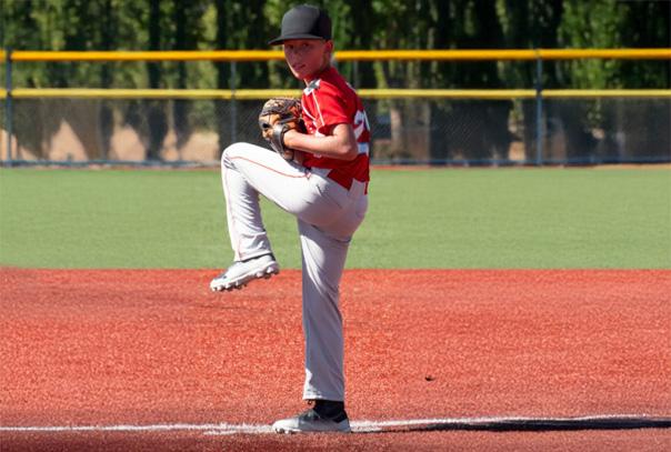 A youth baseball pitcher with a red jersey and black hat at balance point on a turf field