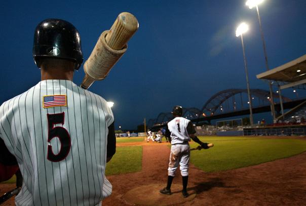 Batter in #5 pinstripe white jersey standing on deck holding a bat with a weight on it looking out towards the field.