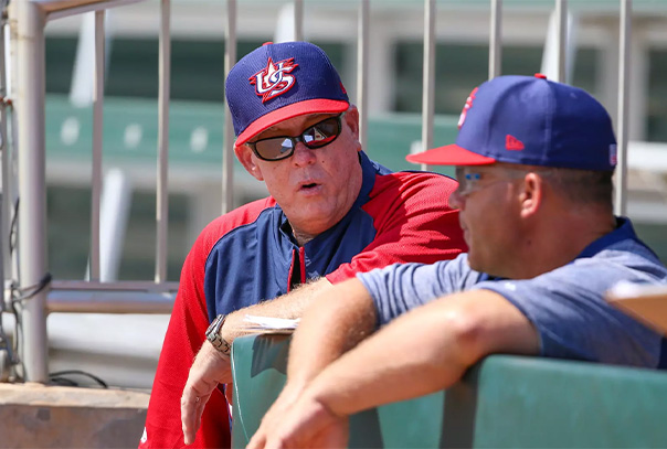 John McCormack in Team USA uniform leaning against dugout rail and talking to another coach in same uniform.