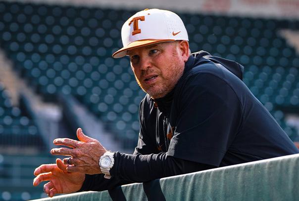 Chuck Box in white University of Texas hat leaning on dugout railing talking